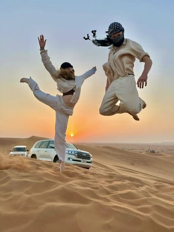 Two guests jumping on a dune beside white 4x4 vehicles