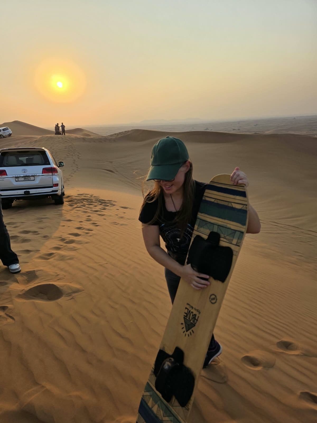 Guest holding a sandboard on a dune with a 4x4 behind her
