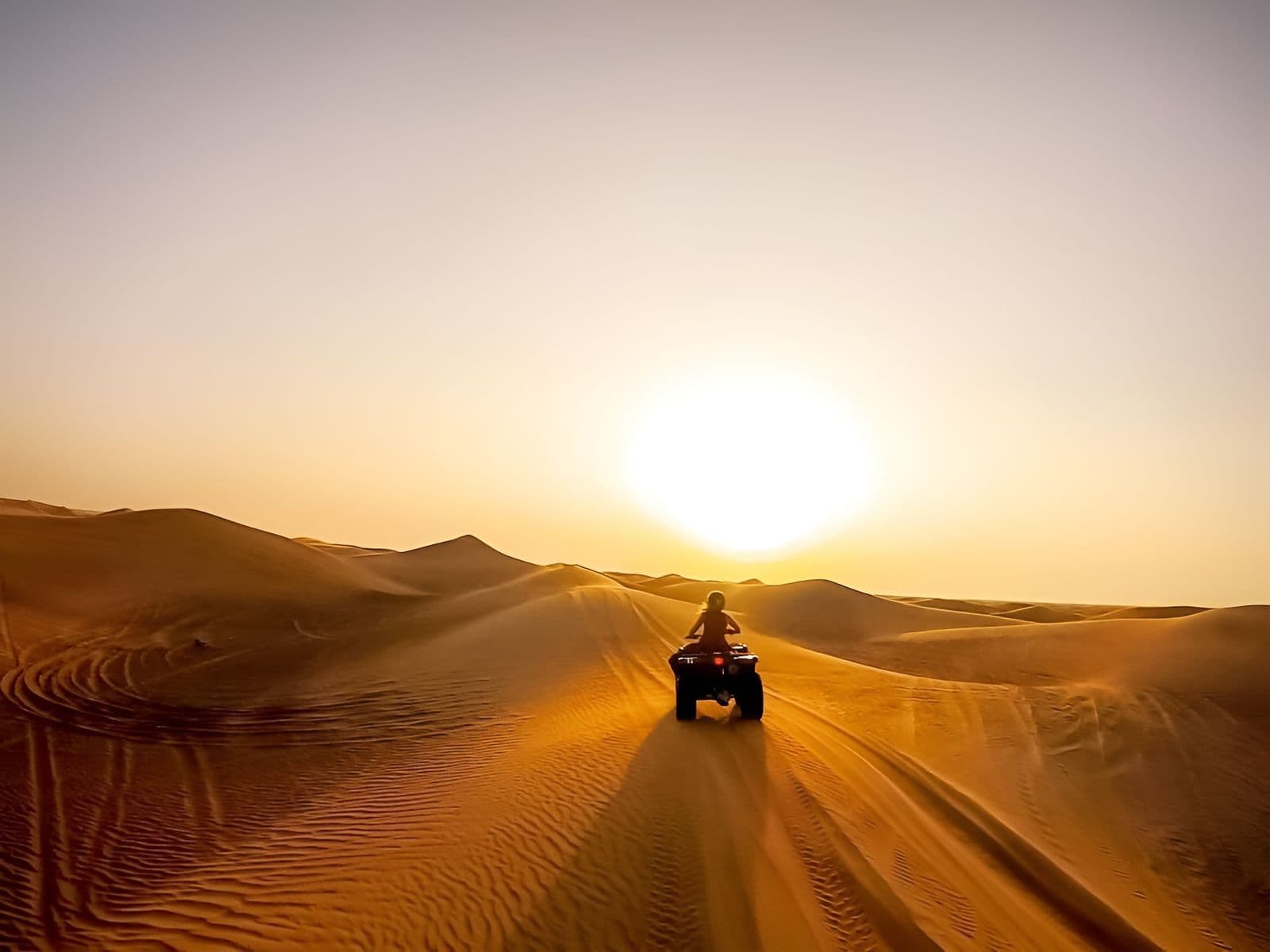 Quad bike rider heading across golden dunes towards the low sun