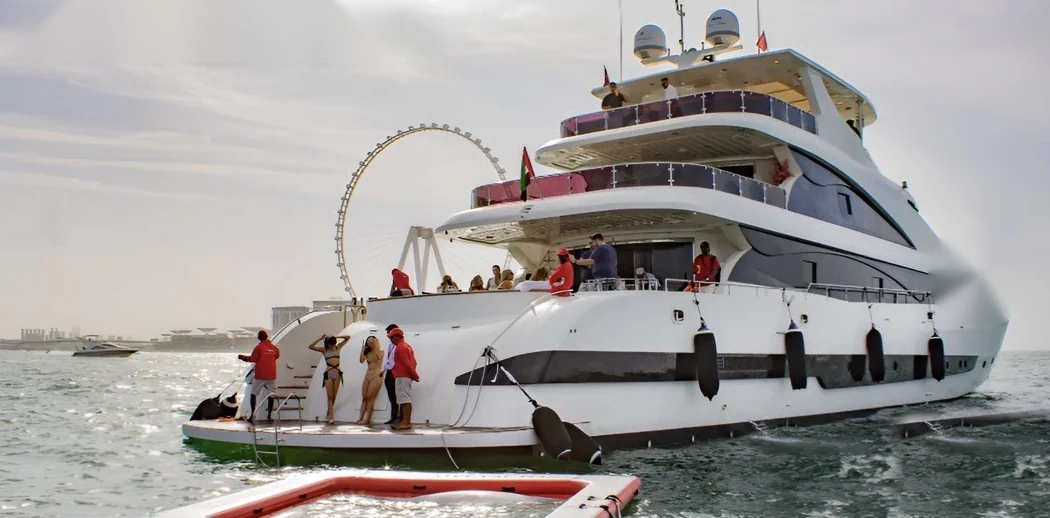 White tri-deck superyacht with guests on the swim deck and Ain Dubai in the background